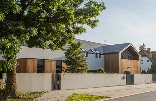 Widok od ulicy na dom w Bielanach Wrocławskich za niskim otynkowanym ogrodzeniem, z drewnianą elewacją parteru i szarym dachem ceramicznym w cieniu rozłożystego drzewa.