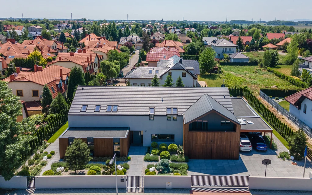 Zdjęcie lotnicze domu w Bielanach Wrocławskich pośród osiedla domów jednorodzinnych z czerwonymi dachami, z widoczną działką, ogrodem i podjazdem z zaparkowanymi samochodami.