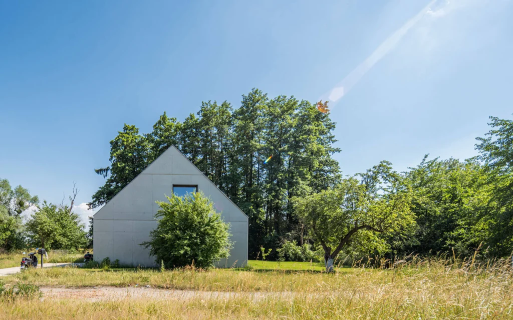Szczytowa ściana domu z betonu architektonicznego z pojedynczym oknem, otoczona łąką i krzewami na tle wysokich drzew nadodrzańskiego łęgu.