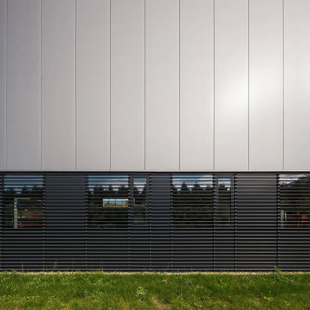 Detal elewacji hali Tornos — pas ciemnych żaluzji aluminiowych osłaniających okna nad zielonym trawnikiem, powyżej gładkie srebrne panele.