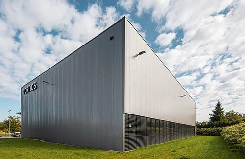 Hala montażowa Tornos widziana od narożnika — kontrastowe srebrne i grafitowe panele elewacyjne, czarny cokół z żaluzjami i podjazd z zaparkowanym samochodem pod pochmurnym niebem.