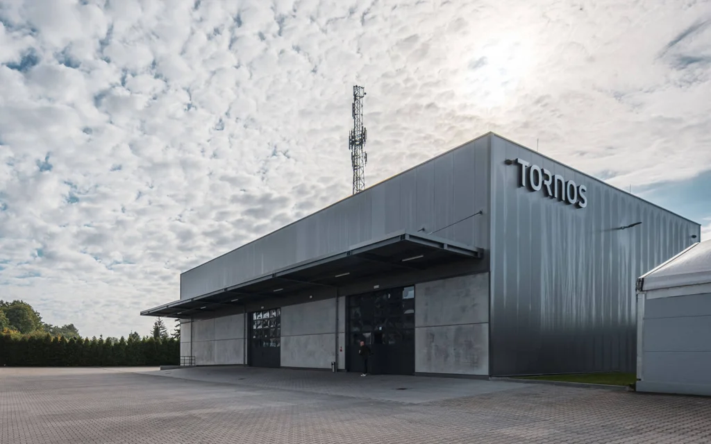 Wejście do hali Tornos osłonięte stalowym zadaszeniem nad betonowymi płytami elewacyjnymi, przeszklone drzwi i idąca osoba, w tle parking i drzewa.