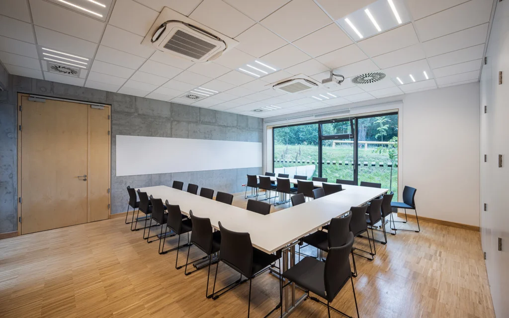 Sala konferencyjna z długim białym stołem ustawionym w podkowę, czarnymi krzesłami, tablicą suchościeralną na betonowej ścianie i widokiem na zielony teren za oknem.