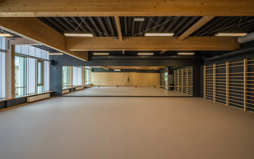 Sala tańca z lustrzaną ścianą, drewnianym drążkiem baletowym, drabinkami gimnastycznymi przy ścianie i widocznym w odbiciu statywem kamery.