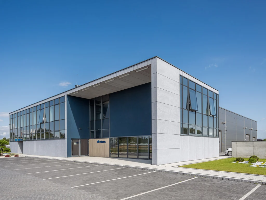 Narożnik budynku łączący przeszkloną część biurową z halą produkcyjną — granatowa wnęka wejściowa, logo Inbras i skwer z niską zielenią.