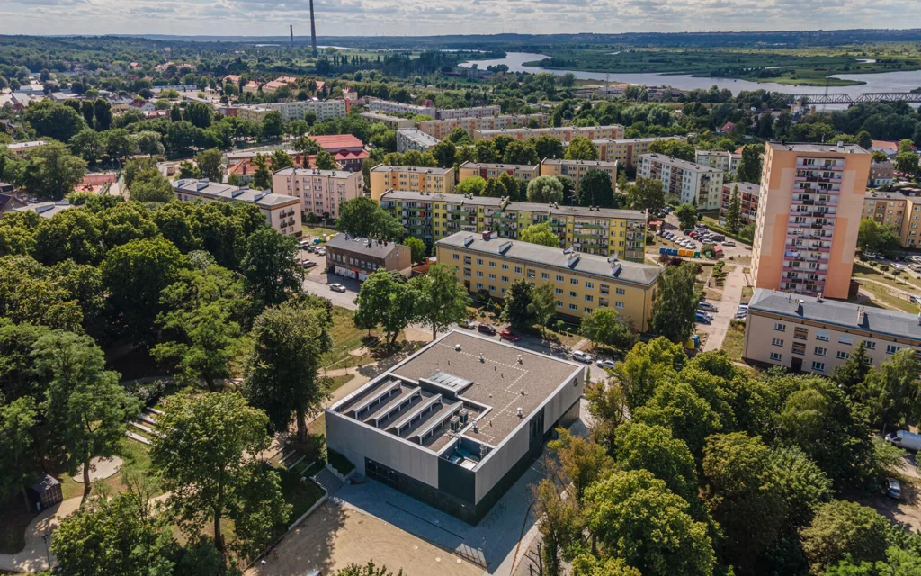Zdjęcie lotnicze pokazujące budynek Domu Kultury pośród zieleni parku na tle kolorowych bloków mieszkalnych osiedla Podjuchy i zakola rzeki w oddali.