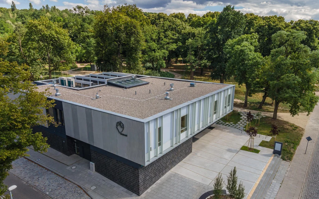 Zdjęcie lotnicze budynku z góry — płaski dach żwirowy ze świetlikami i wentylatorami, logo na attyce, ciemna cokołowa okładzina z klinkieru i otaczający park.