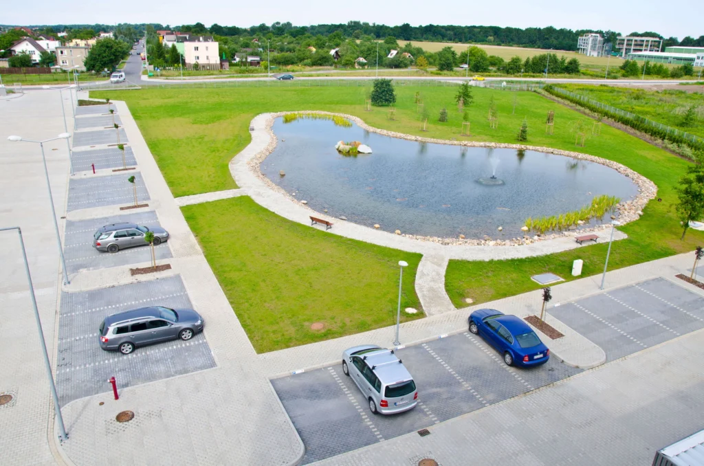 Widok z góry na parking z zadrzewionymi wyspami, zaparkowanymi samochodami i owalnym stawem rekreacyjnym z fontanną i kamienistym brzegiem pośród trawnika.