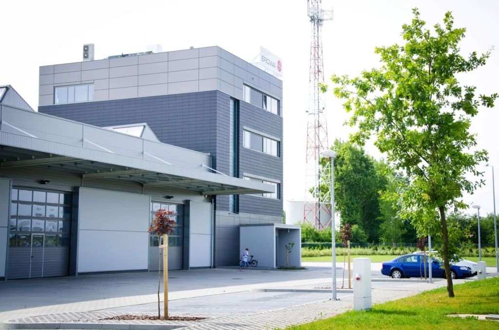 Narożny widok biurowca Erowa w Kątach Wrocławskich — ciemna, wentylowana okładzina ceramiczna czterokondygnacyjnej wieży nad szarą halą montażową, maszt telekomunikacyjny i zaparkowany niebieski samochód w tle.