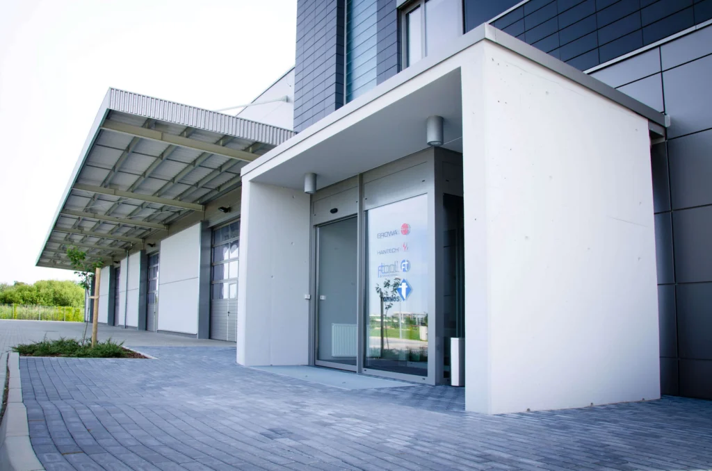 Zadaszone wejście do biurowca z betonowym okapem wspartym na fasadzie oraz przeszklonymi drzwiami z logotypami kilku firm działających w budynku.