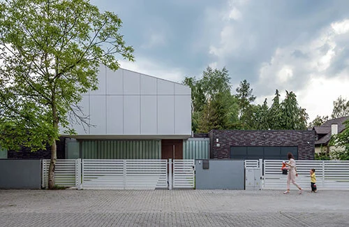 Dom na Krzykach widziany od ulicy — biała geometryczna bryła z ukośnym dachem, ciemna ceglana część garażowa i biała ażurowa brama, przechodząca obok matka z dzieckiem.