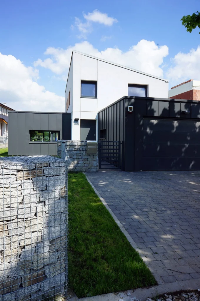 Widok od podjazdu na dom z białym piętrem, ciemnym garażem obłożonym blachą, gabionowym ogrodzeniem i kostką brukową prowadzącą do wejścia.