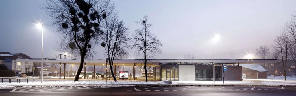 Zimowy widok dworca o zmierzchu — ośnieżony plac, nagie drzewa otaczające przeszklony pawilon pod smukłym dachem, autobusy przy peronach.