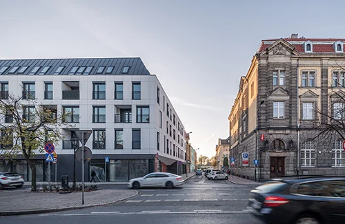 Elewacja frontowa budynku przy Skarbowej w Lesznie o zmierzchu — biała okładzina elewacyjna z czarną stolarką okienną, usługowy parter i sąsiadująca zabytkowa kamienica w tle.