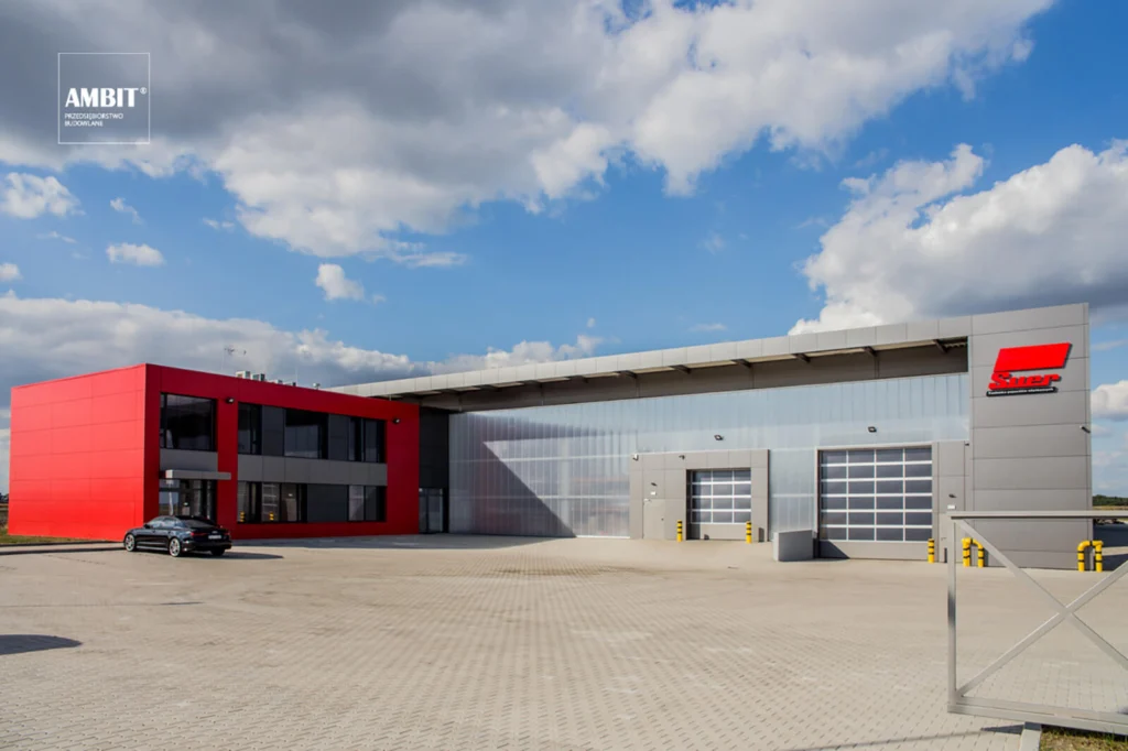 Widok całej hali produkcyjno-magazynowej Suer Polska w Widziszewie — czerwony blok biurowy połączony z szarą halą krytą blachą, zaparkowany czarny samochód na placu.