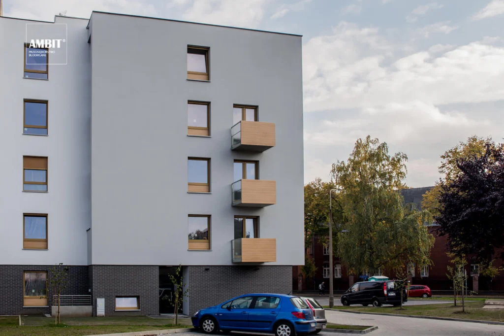 Widok budynku od strony ulicy jesienią, z zaparkowanymi samochodami na pierwszym planie i pasem balkonów z drewnianymi osłonami wzdłuż narożnika elewacji.