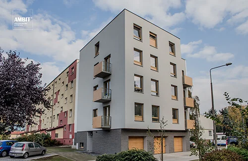 Narożny widok budynku wielorodzinnego przy Ułańskiej w Lesznie — jasna tynkowana bryła z drewnianymi oknami i balkonami, ciemna klinkierowa cegła cokołu z trzema drewnianymi bramami garażowymi.