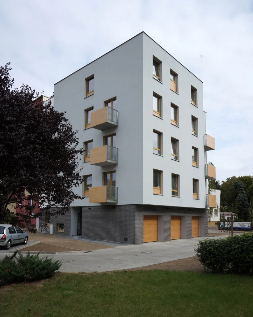 Pionowy kadr narożnika budynku od strony trawnika z drzewem na pierwszym planie — kaskada balkonów osłoniętych drewnianymi panelami nad klinkierową cegłą przyziemia.